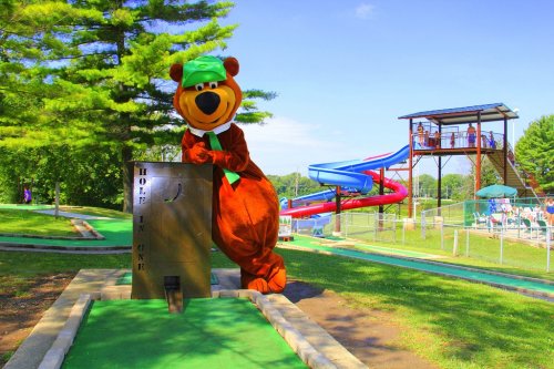 A bear mascot in a green hat leans on a mini-golf hole sign, with a water slide and pool in the background.
