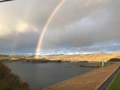 A vibrant rainbow arcs over a calm lake surrounded by autumn-colored hills under a cloudy sky.