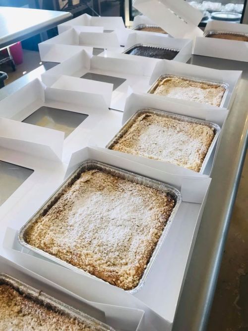 A row of baked goods in aluminum trays, each placed in a white box with a clear window on a countertop.