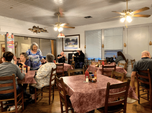 A cozy restaurant interior with patrons at tables, chatting and enjoying their meals under ceiling fans and warm lighting.