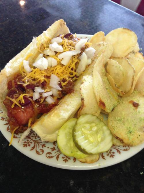 A plate featuring a chili cheese hot dog topped with onions and cheese, alongside crispy potato chips and pickles.