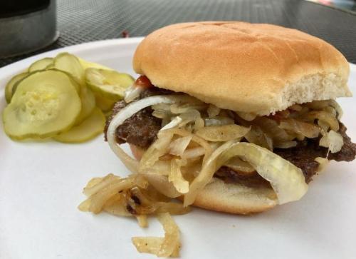A hamburger with sautéed onions and ketchup on a bun, served with a side of pickles on a white plate.