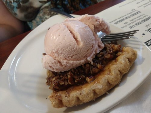 A slice of pecan pie topped with a scoop of pink ice cream on a white plate.