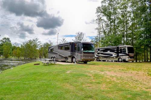 Two RVs parked by a serene lake, surrounded by trees under a partly cloudy sky. Picnic table nearby.