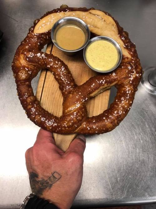 A large, soft pretzel on a wooden board, served with two dipping sauces in metal bowls.
