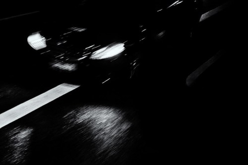 A blurred car driving on a dark road, with reflections of light on the wet surface.