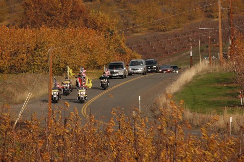 Motorcycles with American flags ride along a winding road, flanked by autumn-colored trees and passing cars.