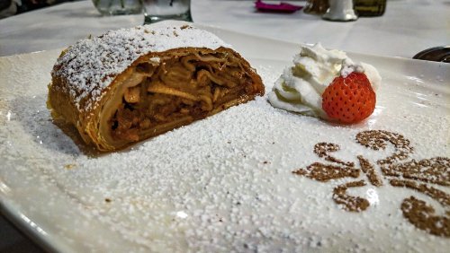A slice of apple strudel dusted with powdered sugar, served with whipped cream and a strawberry on a decorative plate.