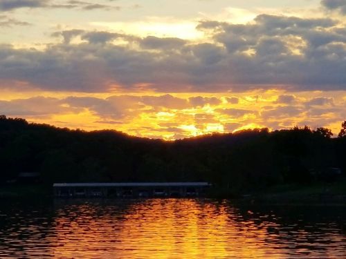 A vibrant sunset over a lake, with orange and yellow hues reflecting on the water and silhouetted trees in the background.