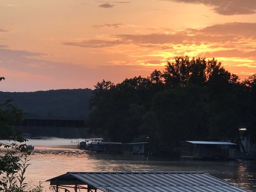 Sunset over a calm river, with silhouettes of trees and boats, and a mist rising from the water.