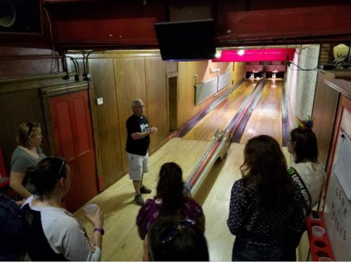 A group of people listens to a speaker in a bowling alley, with lanes visible in the background.