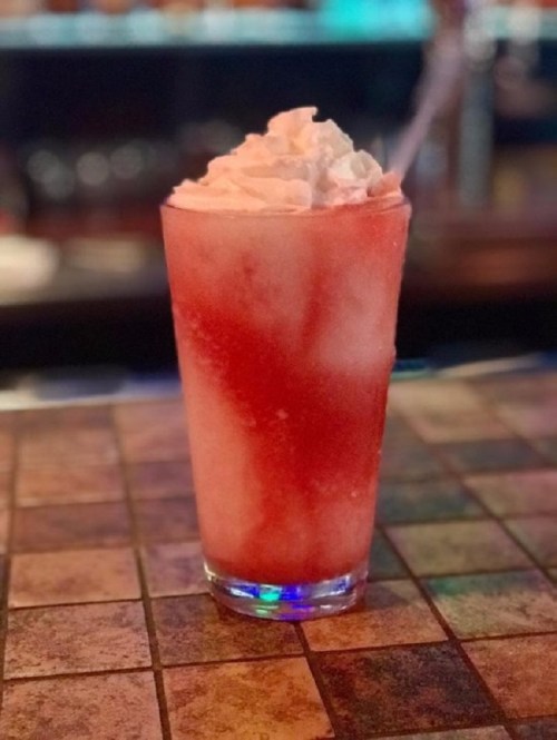 A tall glass filled with a red and white slushy drink topped with whipped cream, sitting on a tiled surface.