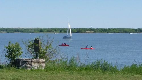 Two kayakers paddle on a calm lake, while a sailboat glides in the background under a clear blue sky.