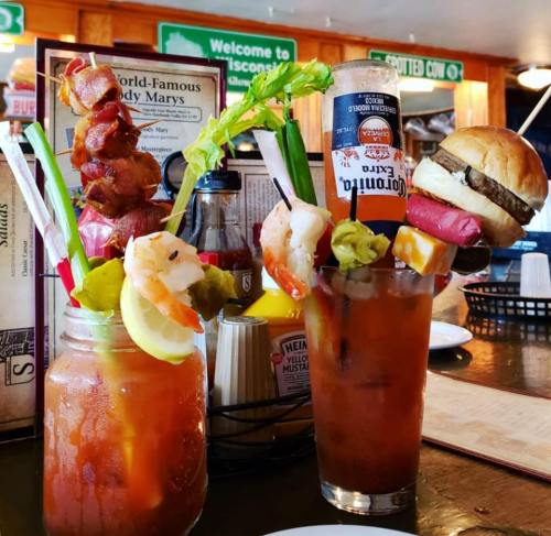 Two elaborate Bloody Mary cocktails with garnishes, including shrimp, celery, and a mini burger, on a restaurant table.