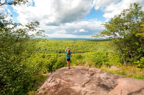 A person stands on a rocky outcrop, overlooking a lush green valley under a partly cloudy sky.