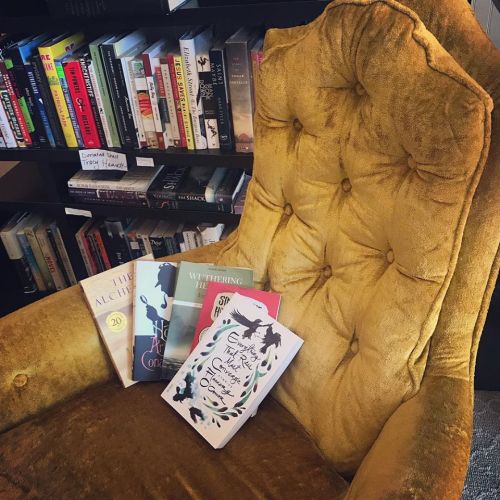 A cozy yellow armchair with several books stacked on it, surrounded by shelves filled with more books.