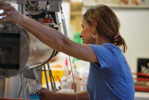 A woman in a blue shirt prepares a drink at a counter, focused on her task in a vibrant café setting.