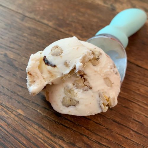 A scoop of ice cream with chunks of nuts, resting on a wooden surface next to a blue ice cream scoop.