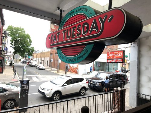 Sign for "Fat Tuesday" restaurant featuring food and frozen drinks, with a view of a street and parked cars.
