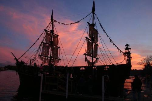 Silhouette of a pirate ship against a colorful sunset sky, with flags and rigging visible.