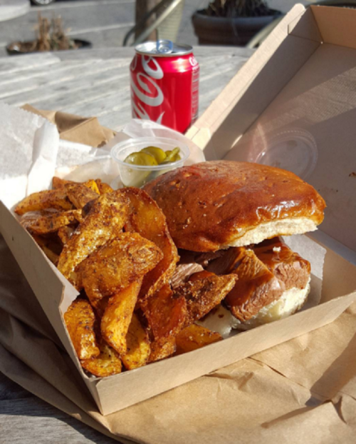 A takeout box with a sandwich, crispy potato wedges, pickles, and a can of soda on a wooden table.