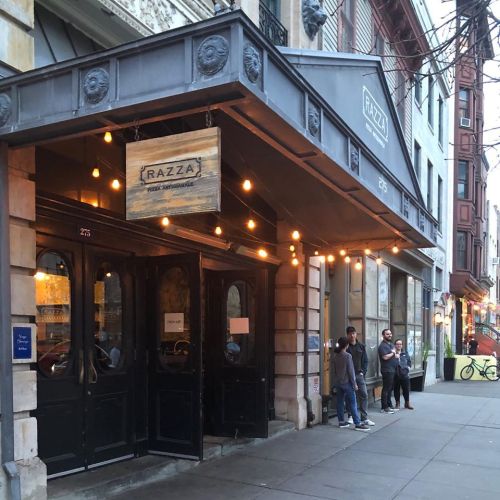 Exterior of Razza restaurant with a wooden sign, string lights, and people standing outside in an urban setting.