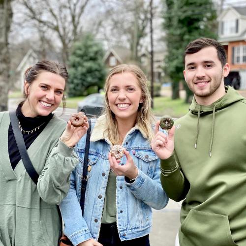 Three friends smile while holding donuts outdoors, dressed in casual clothing against a suburban backdrop.