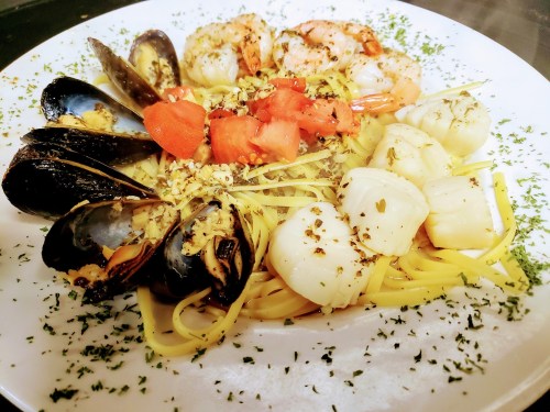 A plate of pasta topped with shrimp, scallops, mussels, and diced tomatoes, garnished with herbs.