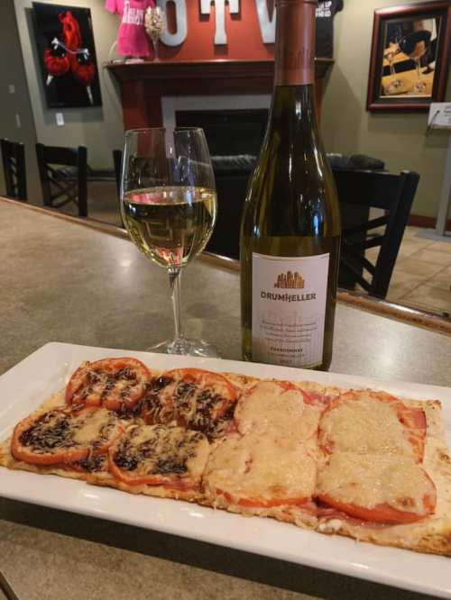 A glass of white wine beside a plate of flatbread topped with melted cheese and sliced tomatoes, set in a cozy restaurant.