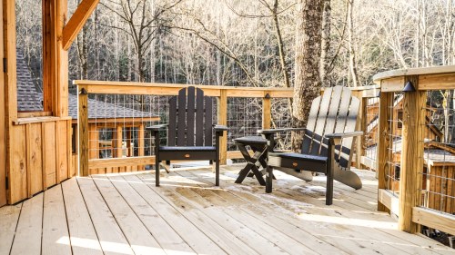 Two black Adirondack chairs and a small table on a wooden deck surrounded by trees in a serene outdoor setting.