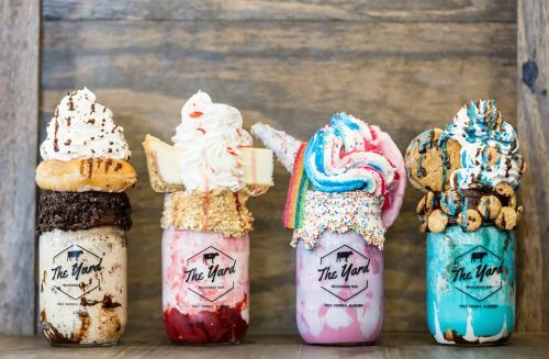 Four colorful milkshakes in jars topped with whipped cream, cookies, donuts, and sprinkles, displayed on a wooden surface.