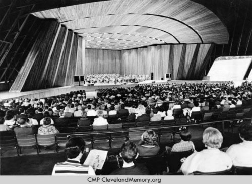 A large audience seated in a modern auditorium with a wooden interior, watching a performance on stage.
