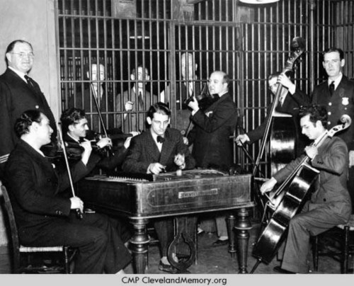 A group of musicians perform in front of prison bars, with several men playing instruments and others watching from behind.