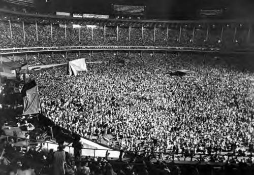 A large crowd fills a stadium, with people gathered closely together under bright lights at night.