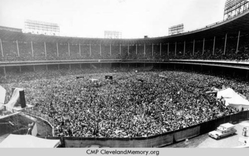 A large crowd fills a stadium, with people gathered for an event, surrounded by tall seating and structures.