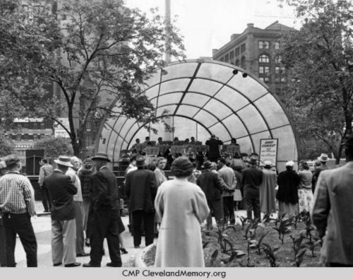 A crowd gathers around a bandstand in a park, with trees and historic buildings in the background.