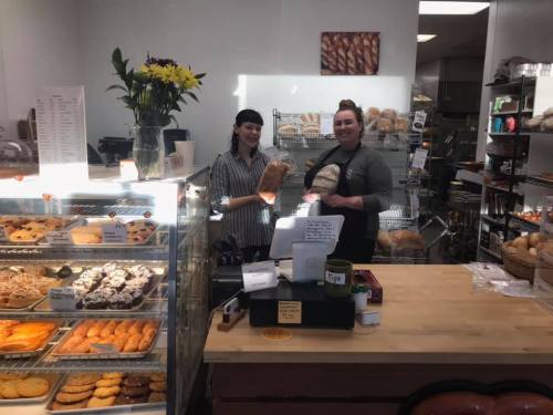 Two bakers smile behind the counter of a bakery, surrounded by fresh bread and pastries. Bright and inviting atmosphere.
