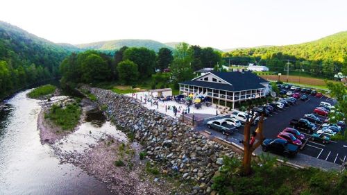 Aerial view of a riverside venue with a parking lot, surrounded by green hills and trees, hosting an outdoor event.