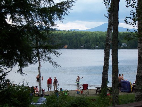 A serene lake scene with people swimming and relaxing on the shore, surrounded by trees and mountains in the background.