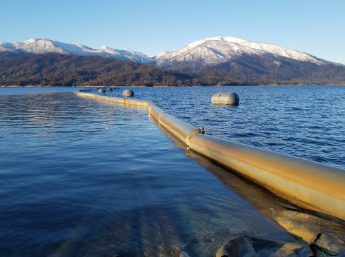 A long pipe extends into a calm lake, with snow-capped mountains in the background under a clear blue sky.