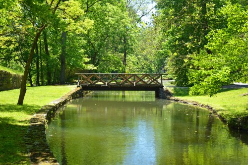 A wooden bridge spans a calm, reflective stream surrounded by lush green trees and grass.
