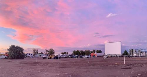 A sunset sky with pink and purple hues over a drive-in movie theater, featuring parked cars and a large blank screen.