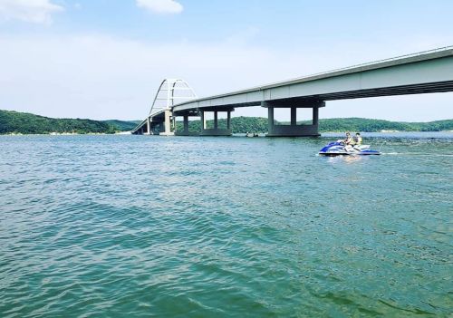 A jet ski glides across a calm lake beneath a large bridge, surrounded by green hills and a clear blue sky.