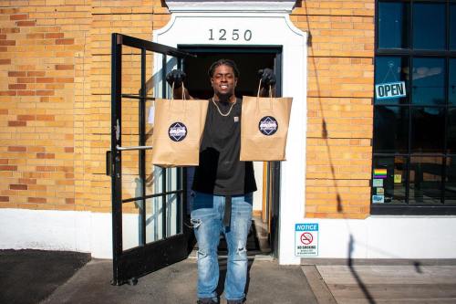 A person stands at an open door holding two paper bags, smiling in front of a brick building.