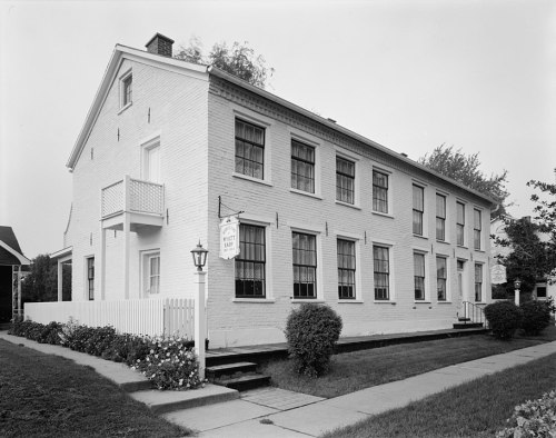 A white brick building with multiple windows, a balcony, and a sign, surrounded by greenery and a sidewalk.