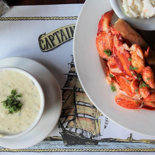 A plate with lobster on a bun, coleslaw, and a bowl of clam chowder on a nautical-themed placemat.