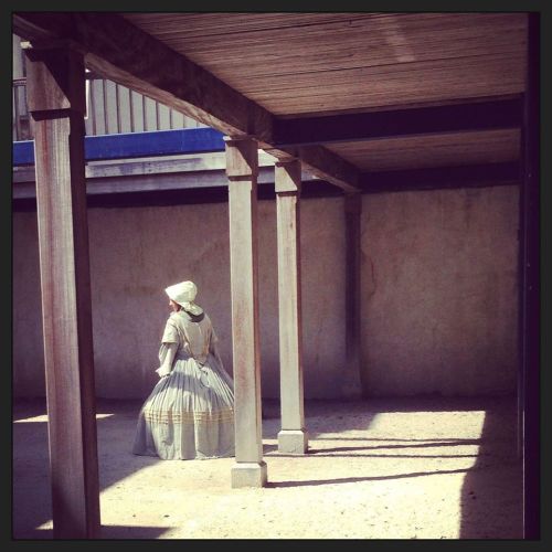 A woman in a historical dress stands under wooden beams in a sunlit, empty courtyard.