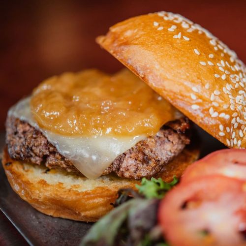 A close-up of a burger with a sesame seed bun, melted cheese, and a slice of meat, accompanied by tomato and greens.
