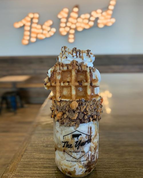 A tall dessert jar filled with ice cream, topped with whipped cream, a waffle, and crushed toppings, against a lit sign.