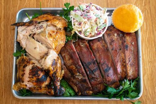 A metal tray filled with grilled chicken, ribs, coleslaw, and a cornbread muffin, garnished with parsley.